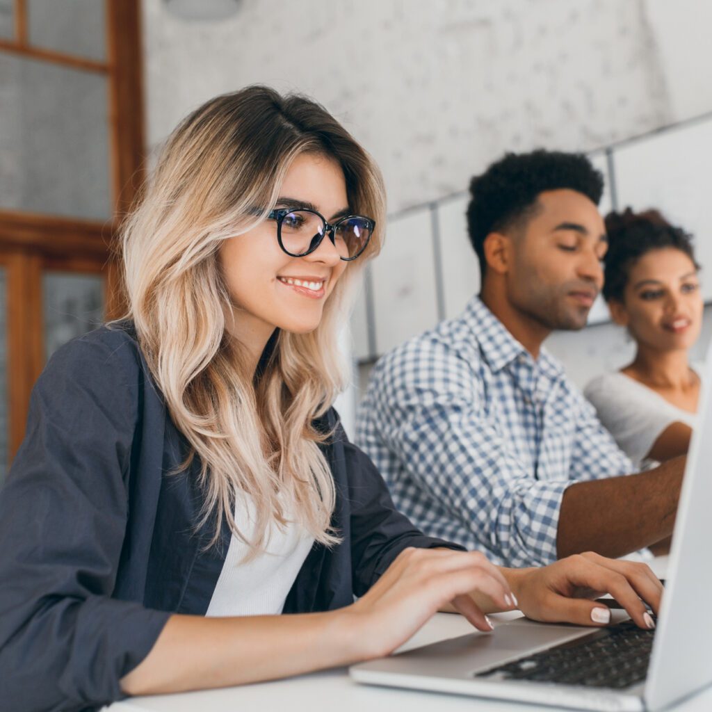 beautiful-curly-female-freelancer-with-cute-manicure-using-laptop-and-smiling-indoor-portrait-of-blonde-secretary-sitting-beside-african-coworker-in-blue-shirt